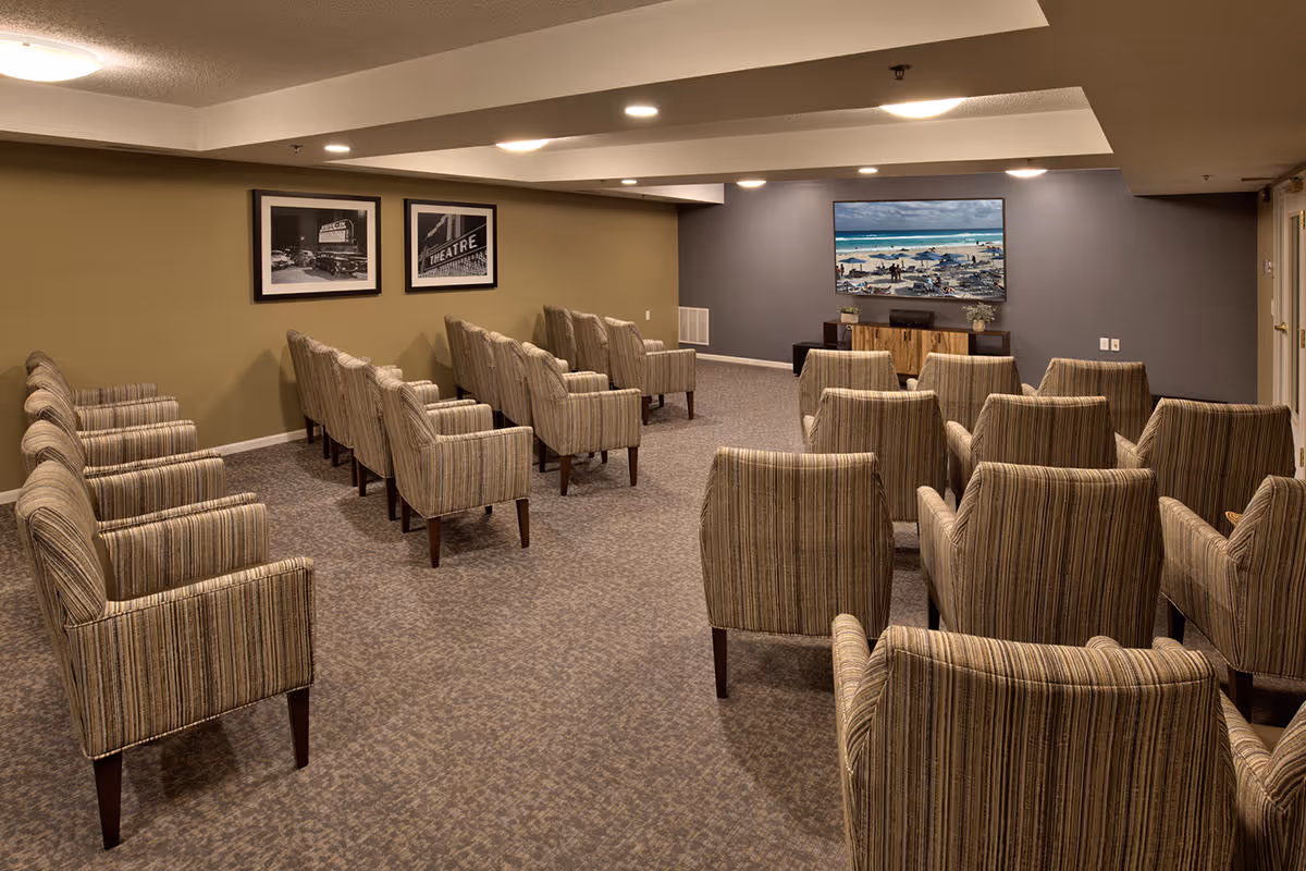 A small theater room with multiple rows of striped upholstered chairs facing a large flat-screen TV mounted on a gray wall. The room has beige walls, carpeted floor, and two black-and-white framed pictures on one side wall.