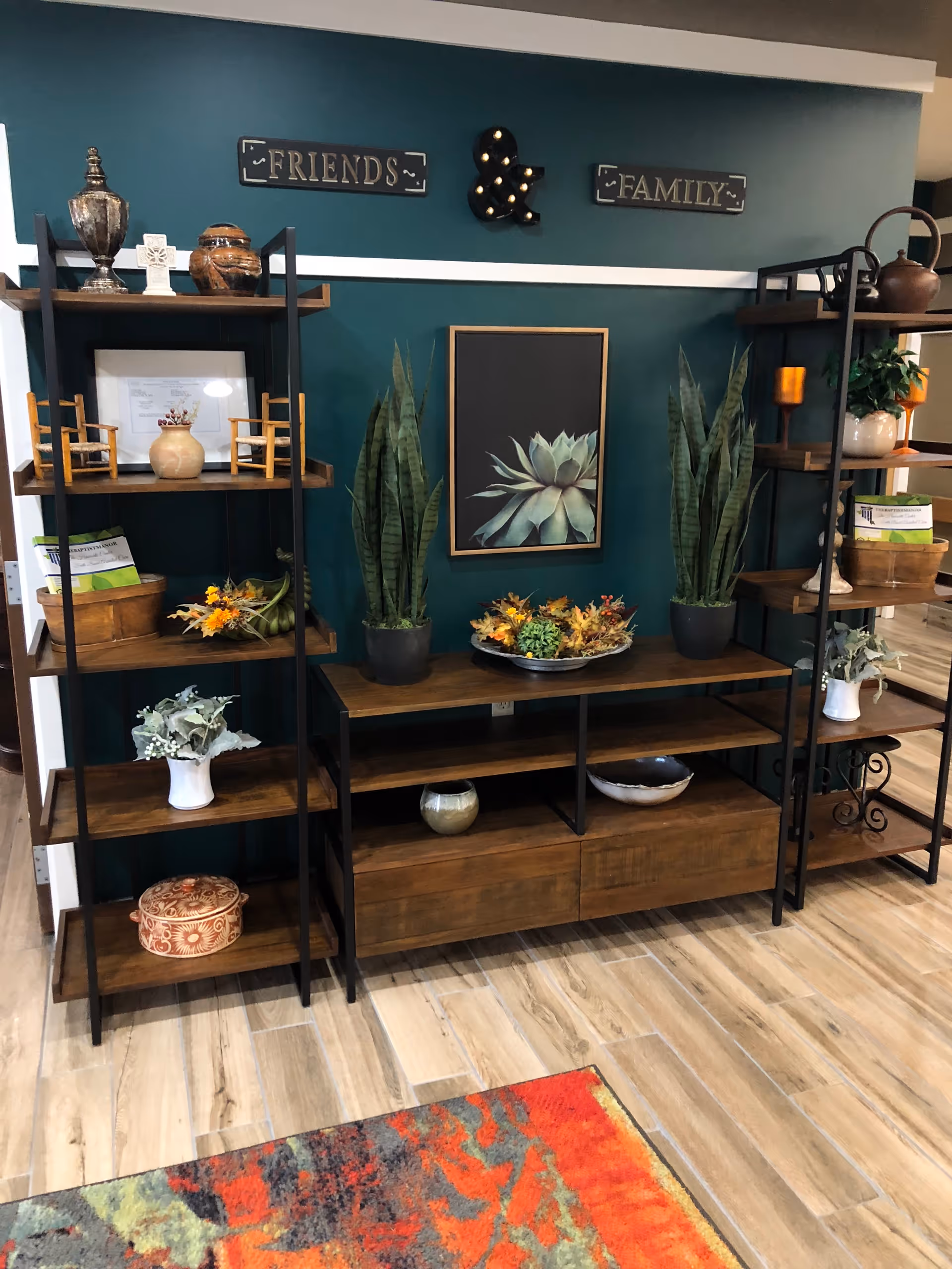 A decorative shelving unit against a teal wall with wooden shelves holding various decorative items including plants, pottery, small chairs, and books. Above the shelves are three wall decorations: a sign that says 'FRIENDS', a lit ampersand symbol, and a sign that says 'FAMILY'. The floor is wooden with a colorful rug partially visible in the foreground.
