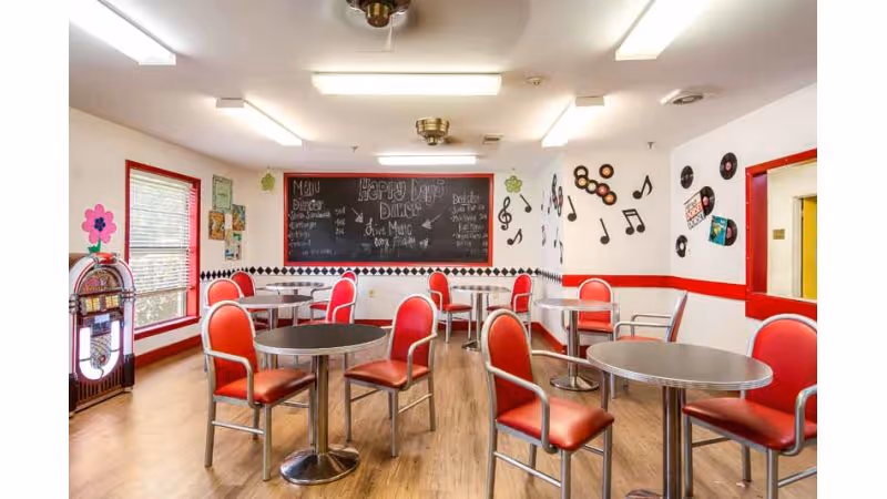 A bright dining room with red and silver chairs around round tables on a wooden floor. The walls are decorated with musical notes, records, and a chalkboard menu that reads 'Happy Days Diner' with food and drink options. A jukebox is visible near a window with red trim.