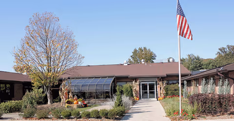 Exterior view of The Oaks at Northpointe facility showing a single-story building with a brown roof, a glass entrance, landscaped garden with shrubs and flowers, a large tree with autumn leaves, and an American flag on a flagpole in front.