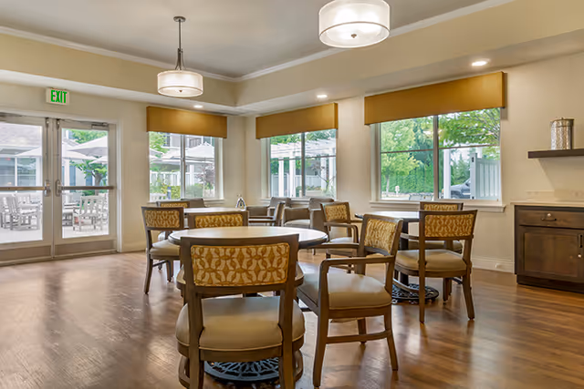 A bright dining room with round wooden tables and cushioned chairs featuring patterned backs. Large windows with yellow valances let in natural light and offer a view of an outdoor patio area with umbrellas. The room has wooden flooring, light-colored walls, and modern ceiling lights.