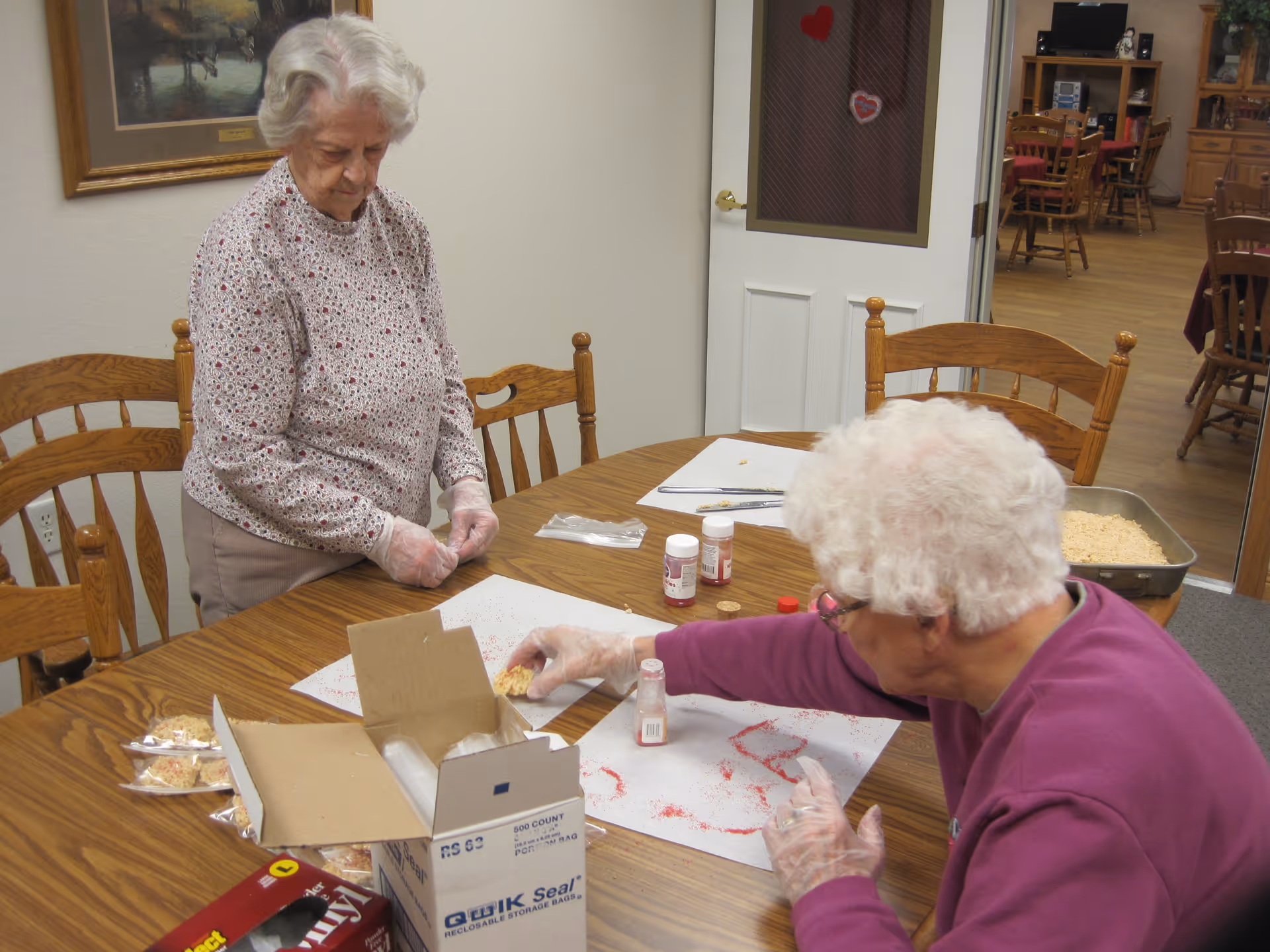 Two elderly women sitting at a wooden table in a room, engaging in a craft or baking activity. One woman is standing and looking down at the table, while the other is seated and reaching for an item on the table. The table has various supplies including a box of resealable storage bags, small containers, and trays with food items. The room has wooden chairs, a door with heart decorations, and a dining area visible in the background.