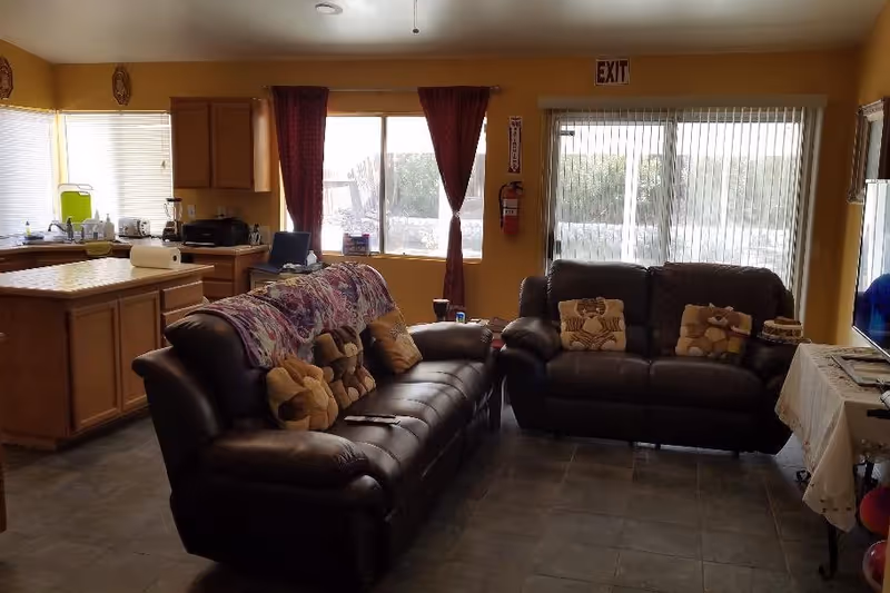 Interior view of a senior care facility living room and kitchen area. The living room has two dark leather sofas with patterned cushions and a floral throw blanket. The kitchen features wooden cabinets, a countertop with a paper towel roll, and various kitchen appliances. There are windows with red curtains and vertical blinds, and an exit sign above a sliding glass door.