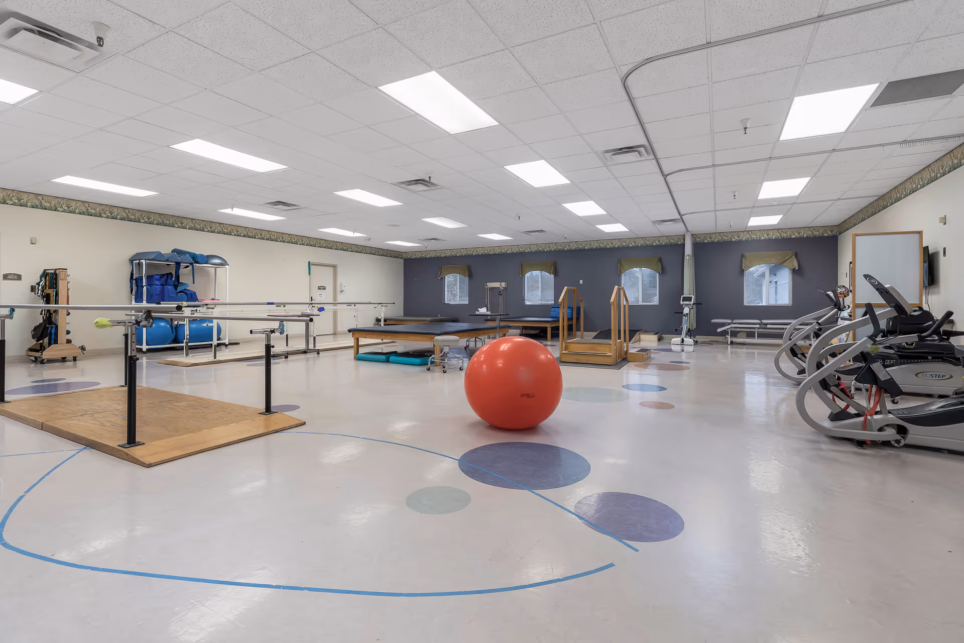 A spacious physical therapy or rehabilitation room with exercise equipment including parallel bars, a large red exercise ball, treadmills, therapy tables, and various other exercise tools. The room has a light-colored floor with circular patterns, a drop ceiling with fluorescent lights, and windows with valances on the far wall.