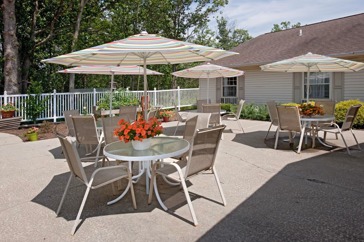 Outdoor patio area with several round glass tables, each surrounded by four beige mesh chairs. Each table has a large striped umbrella providing shade and a flower pot with red flowers as a centerpiece. The patio is adjacent to a light-colored building with windows and surrounded by greenery and a white fence.