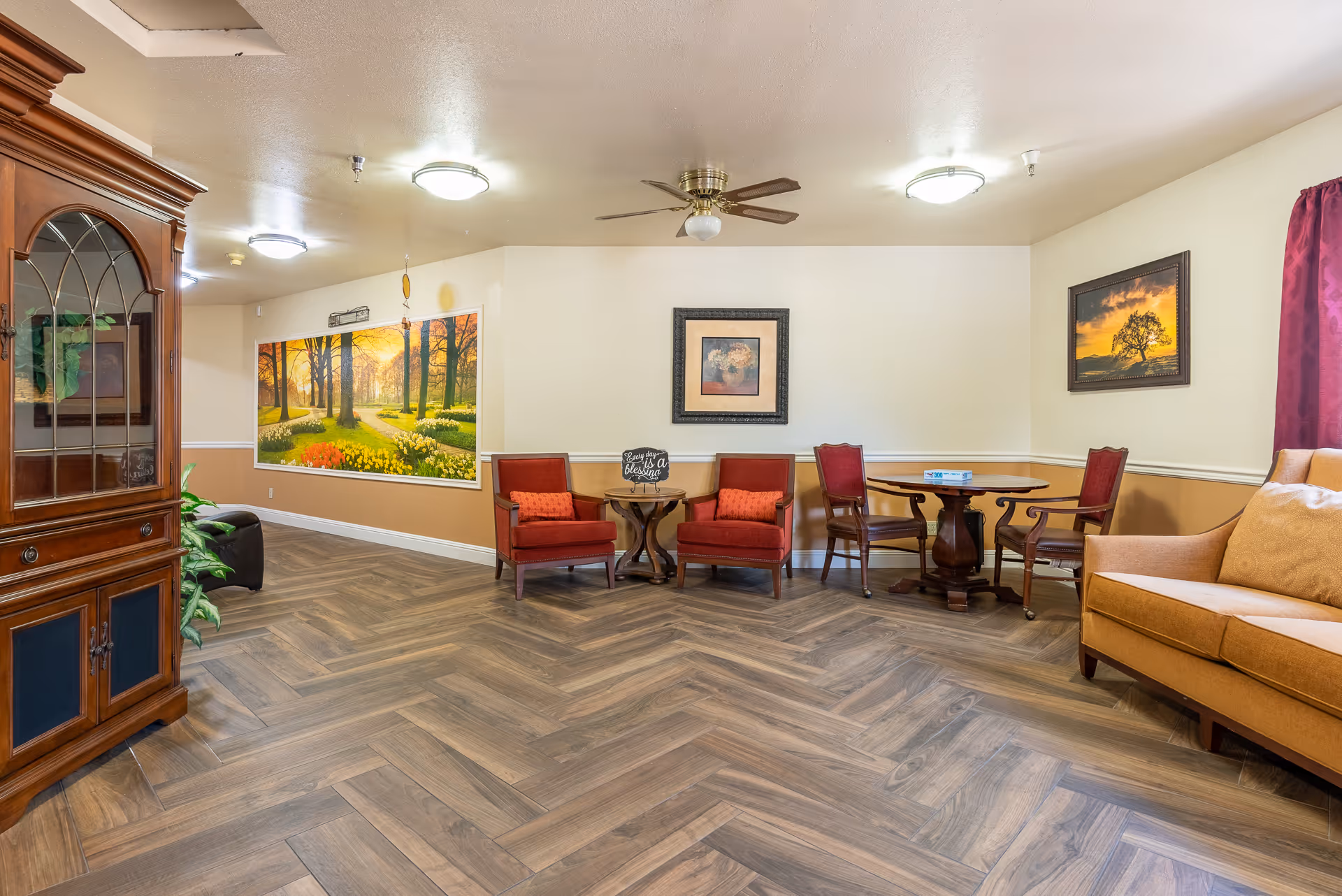 A cozy common area in Valley Crest Memory Care featuring wooden herringbone flooring, two red armchairs with orange cushions around a small round wooden table with a sign that reads 'Every day is a blessing', a round wooden table with two wooden chairs, a mustard yellow sofa with matching cushions, framed artwork on the walls, a ceiling fan, and a large mural of a sunlit forest path with flowers.