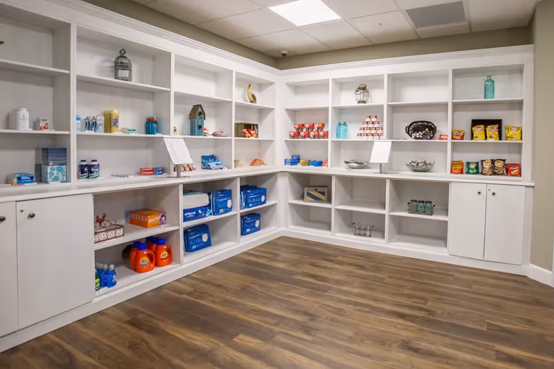 A corner of a room with white built-in shelves stocked with various items including snacks, canned goods, bottled drinks, laundry detergent, and personal care products. The room has wood flooring and a light-colored wall.