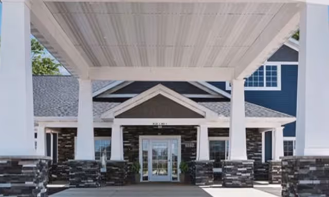 Entrance of a building with a covered driveway supported by white columns with stone bases, leading to glass double doors under a peaked roof section.