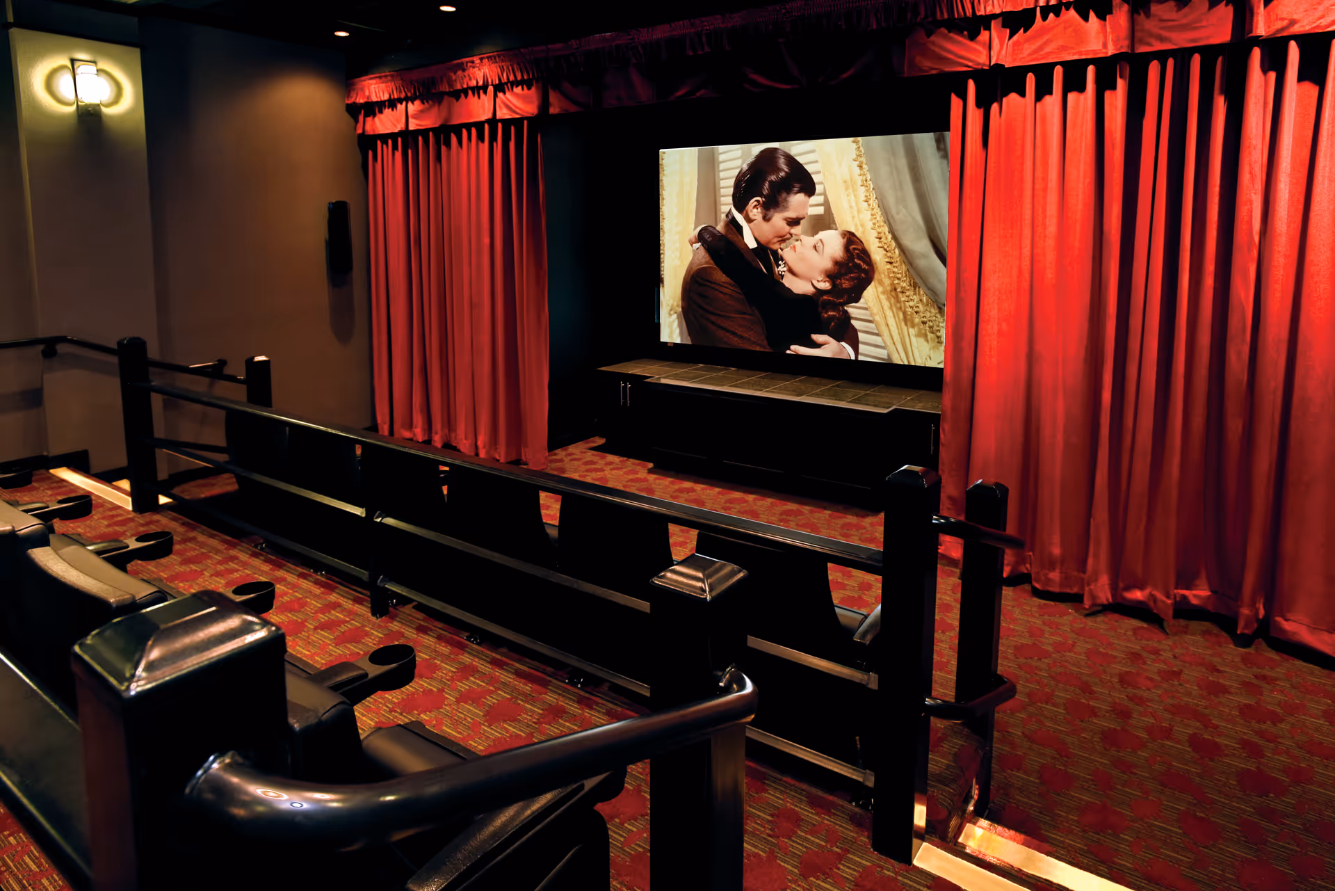 Interior view of a small movie theater room with red curtains on the sides of the screen, a movie playing showing a man and woman embracing, and rows of black leather seats with armrests and cup holders.