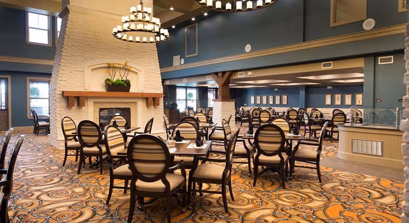 Spacious dining room with multiple round tables and cushioned chairs arranged neatly on a patterned carpet. A large white brick fireplace with a wooden mantel and a decorative plant is centrally located. The room features high ceilings with elegant chandeliers and large windows allowing natural light to enter.