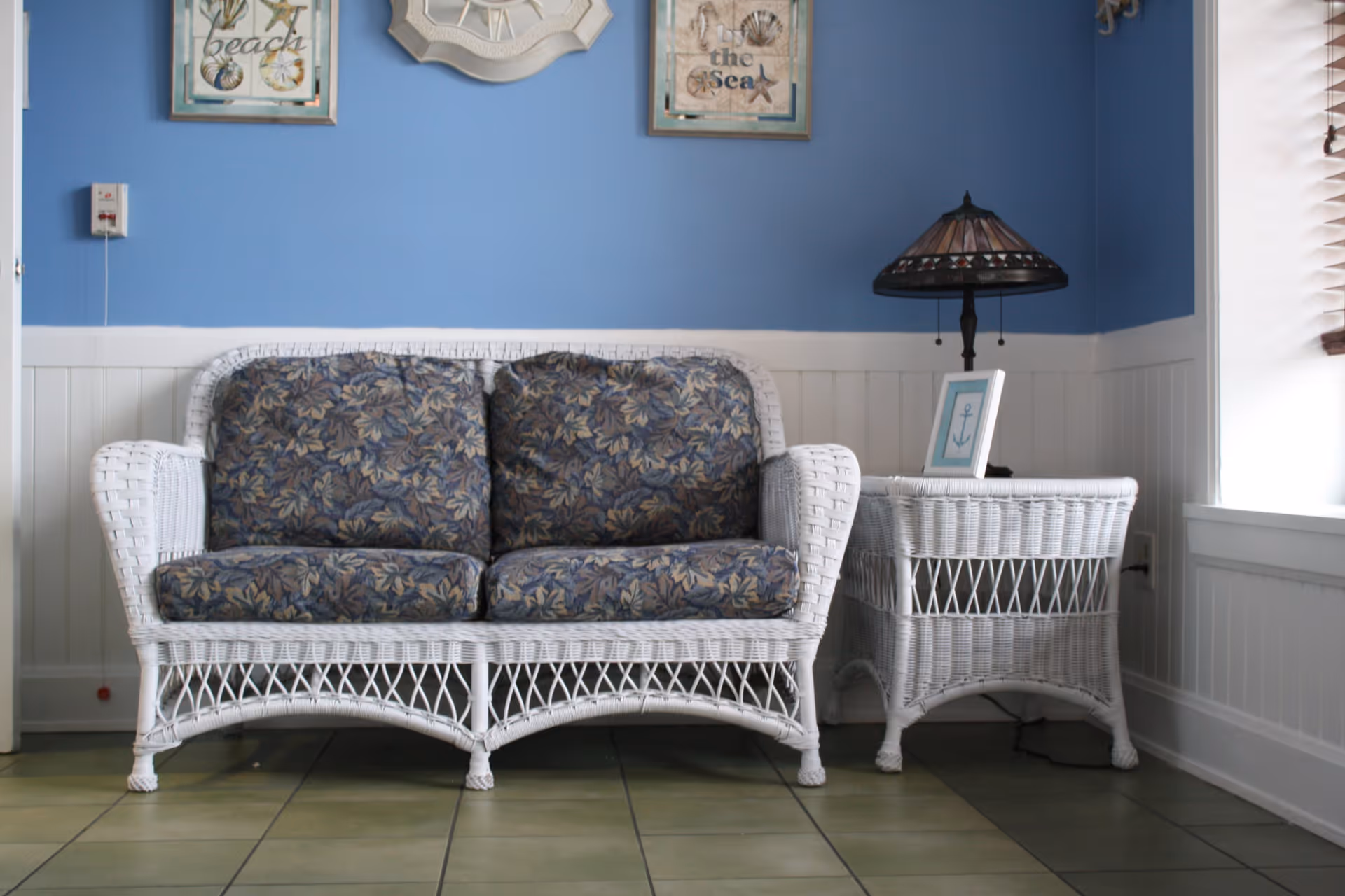 A cozy seating area with a white wicker loveseat featuring floral patterned cushions and a matching wicker side table. On the side table is a small framed picture with an anchor and a decorative lamp with a stained glass shade. The wall behind is painted blue with white wainscoting and decorated with nautical-themed artwork and a ship's wheel.