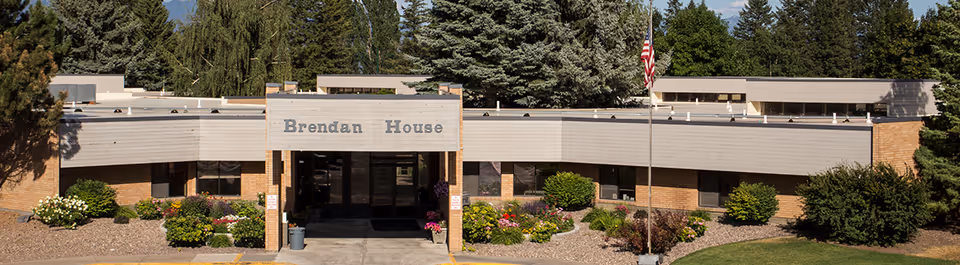 Front entrance of the Brendan House building with a covered entry, landscaped flowerbeds, and an American flag.