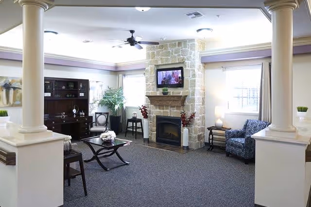 A cozy living room area with a stone fireplace and a mounted flat-screen TV above it. The room features a dark wooden cabinet, a glass coffee table with a flower arrangement, a blue patterned armchair, and a small side table with a lamp. There are two large windows with curtains allowing natural light to enter, and decorative columns framing the space.