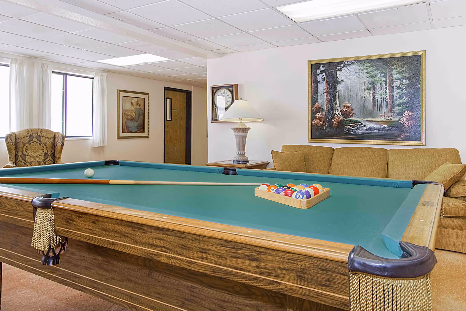Billiards table in a community lounge with couches, an armchair, lamp and framed paintings.