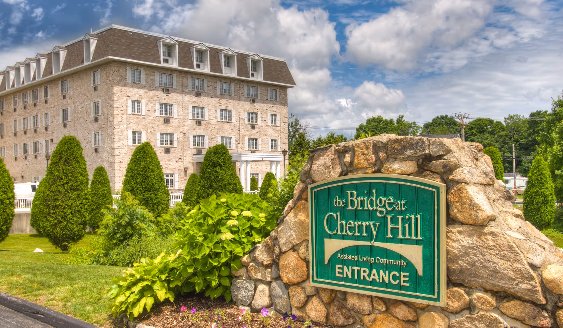 Exterior view of The Bridge at Cherry Hill assisted living community building with a stone sign in the foreground surrounded by greenery and flowers under a partly cloudy sky.