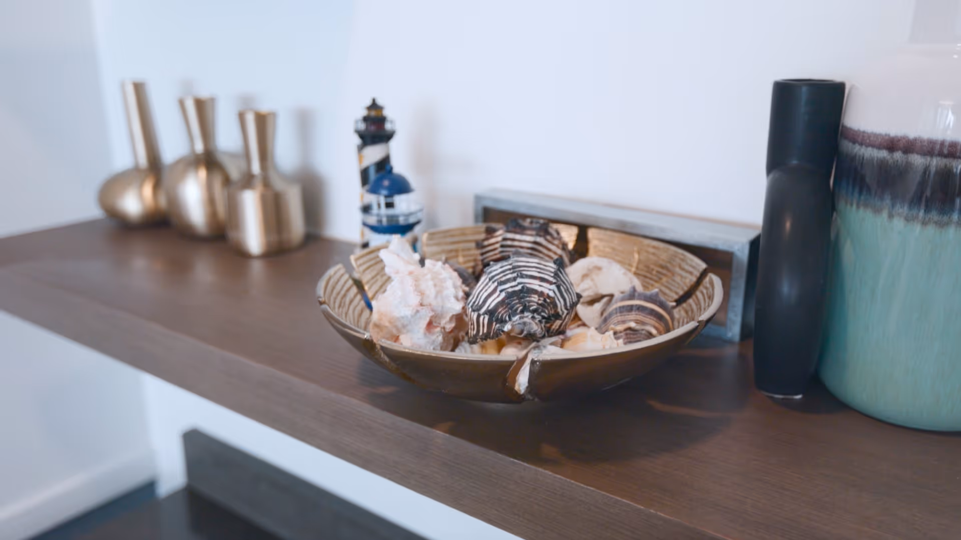 Close-up of a wooden shelf displaying decorative items including a bowl filled with seashells, a small lighthouse figurine, three metallic vases, and two tall vases in black and turquoise colors.