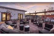 Outdoor patio area at sunset with multiple cushioned chairs and small tables arranged for seating. The space is decorated with string lights overhead and has a view of surrounding buildings and hills in the background.