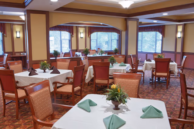 A dining room with multiple tables covered in white tablecloths, each set with green folded napkins and a small floral centerpiece. The room has wooden chairs with patterned upholstery, large windows with blinds, and warm yellow walls accented with wooden trim.