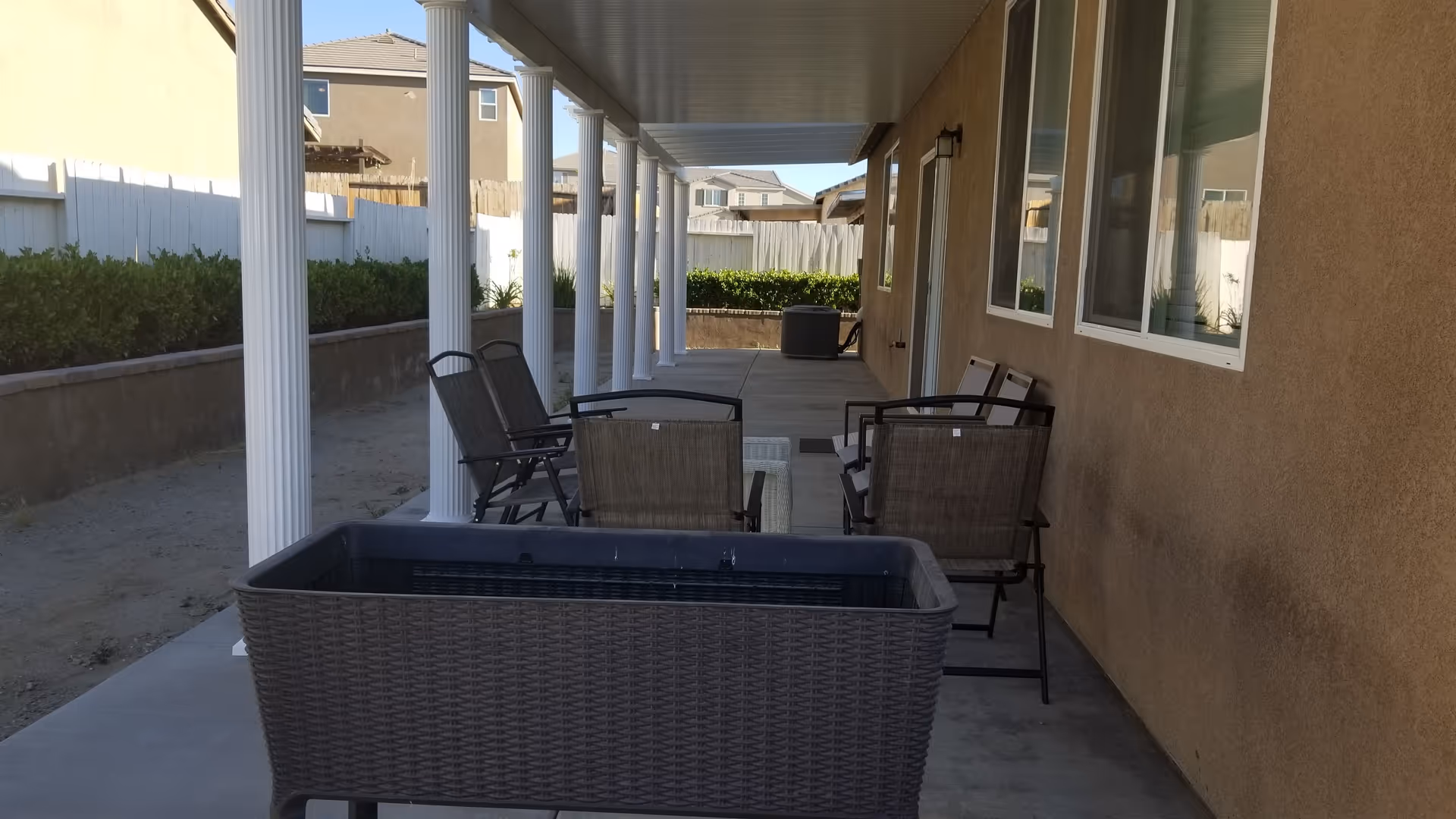 Covered outdoor patio area with several chairs and a small table arranged along the side of a building. White columns support the roof of the patio, and there is a garden bed with green shrubs along a white fence in the background.