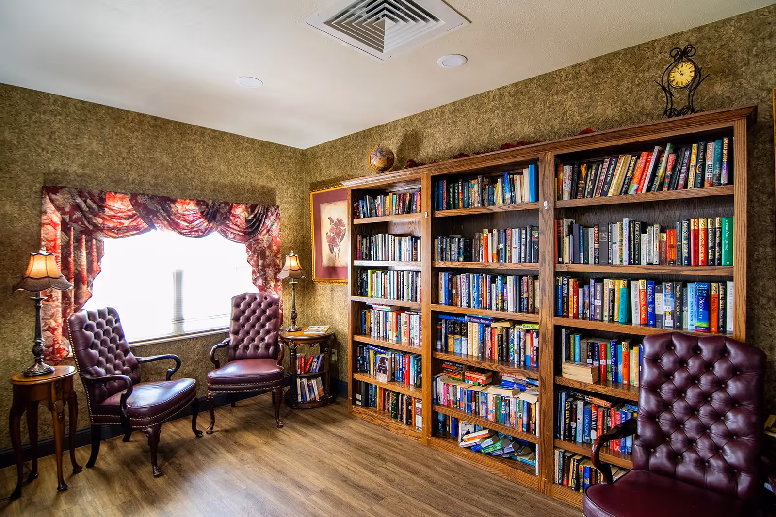 A cozy reading room with three burgundy leather tufted armchairs, two wooden side tables with lamps, a large window with floral valance curtains, and three wooden bookshelves filled with books against a textured beige wall. A framed floral picture and a decorative clock are also visible on the wall.