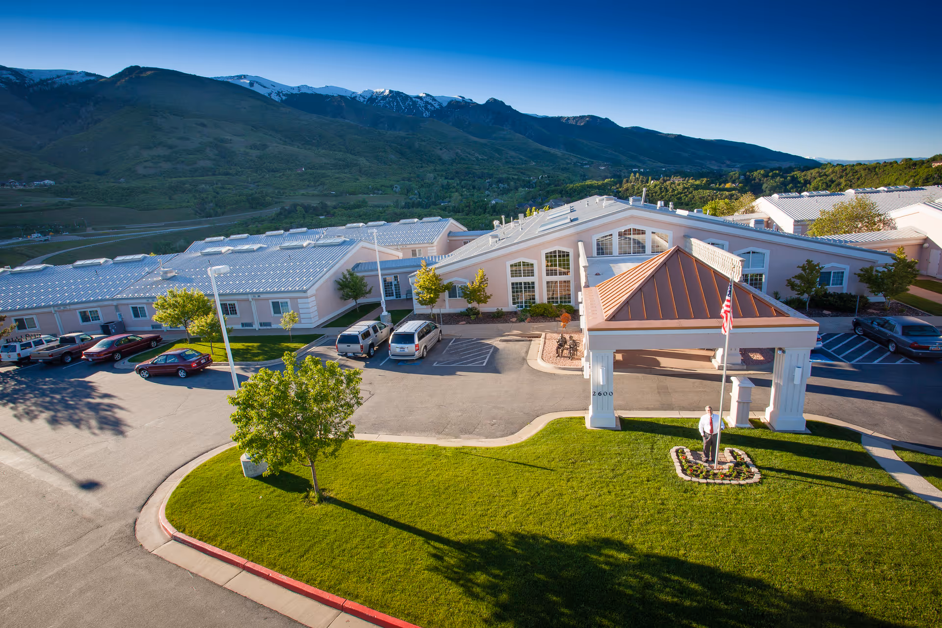 Aerial view of Apple Inn at Apple Village front entrance, with flag, parked cars, and mountains.