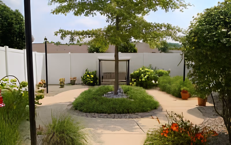 Outdoor garden area with a central tree surrounded by green shrubs and a circular stone border. There are paved walkways leading to a shaded bench swing against a white fence. Various plants and flowers are visible along the paths under a clear sky.