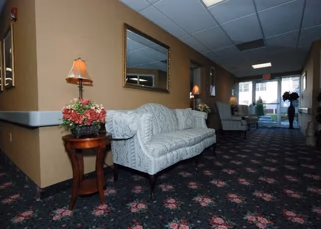 Interior hallway of a nursing and rehabilitation center featuring a patterned carpet with floral designs, a white upholstered sofa, wooden side tables with floral arrangements and lamps, framed mirrors on the walls, and large windows at the far end letting in natural light.