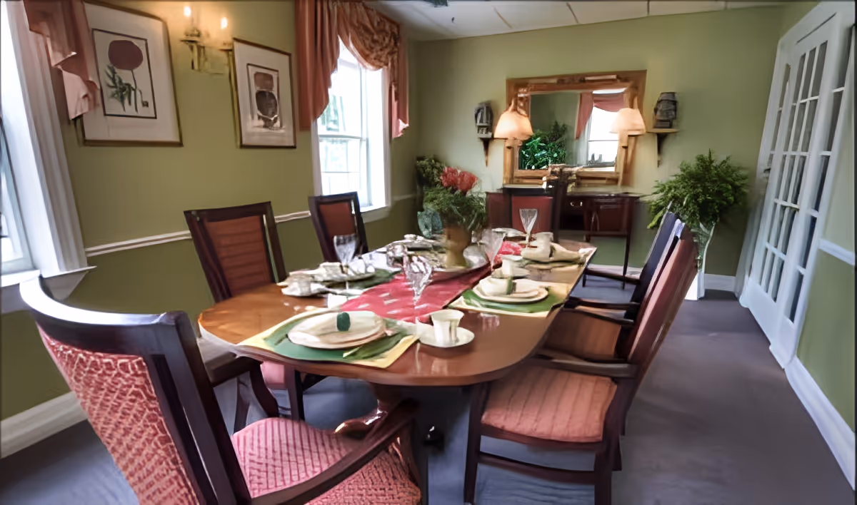 A dining room with a wooden table set for six people, featuring plates, cups, and napkins. The room has green walls, two windows with curtains, framed artwork, a sideboard with a large mirror, and two lamps. There are six wooden chairs with red cushioned seats around the table.