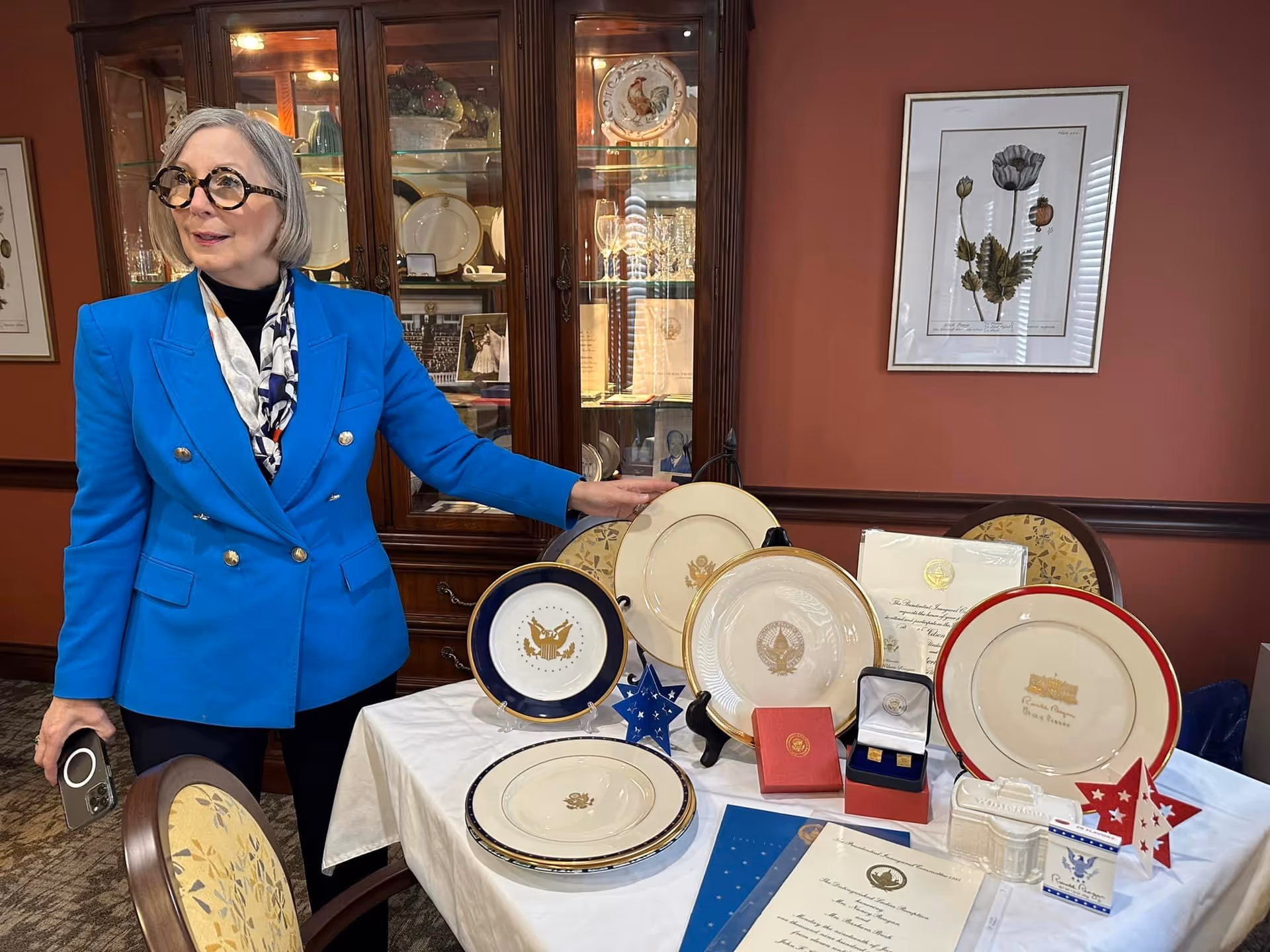 A woman stands beside a table displaying decorative plates, medals, and documents in a dining-room interior with a china cabinet behind her.