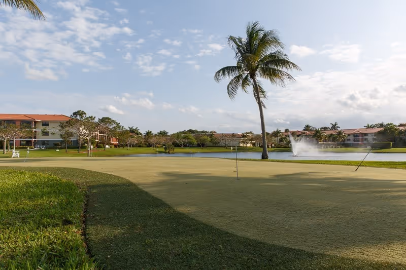 Landscaped exterior with a palm tree, putting green, pond and fountain in front of low-rise residential buildings under a partly cloudy sky.