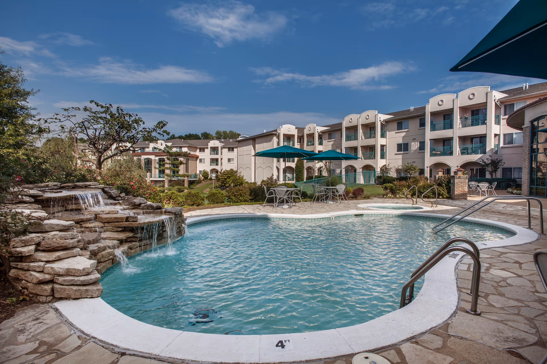 Outdoor swimming pool with a small waterfall feature on the left side, surrounded by a stone deck. Several tables with green umbrellas and chairs are placed near the pool. In the background, there is a multi-story residential building with balconies and landscaped greenery under a partly cloudy blue sky.