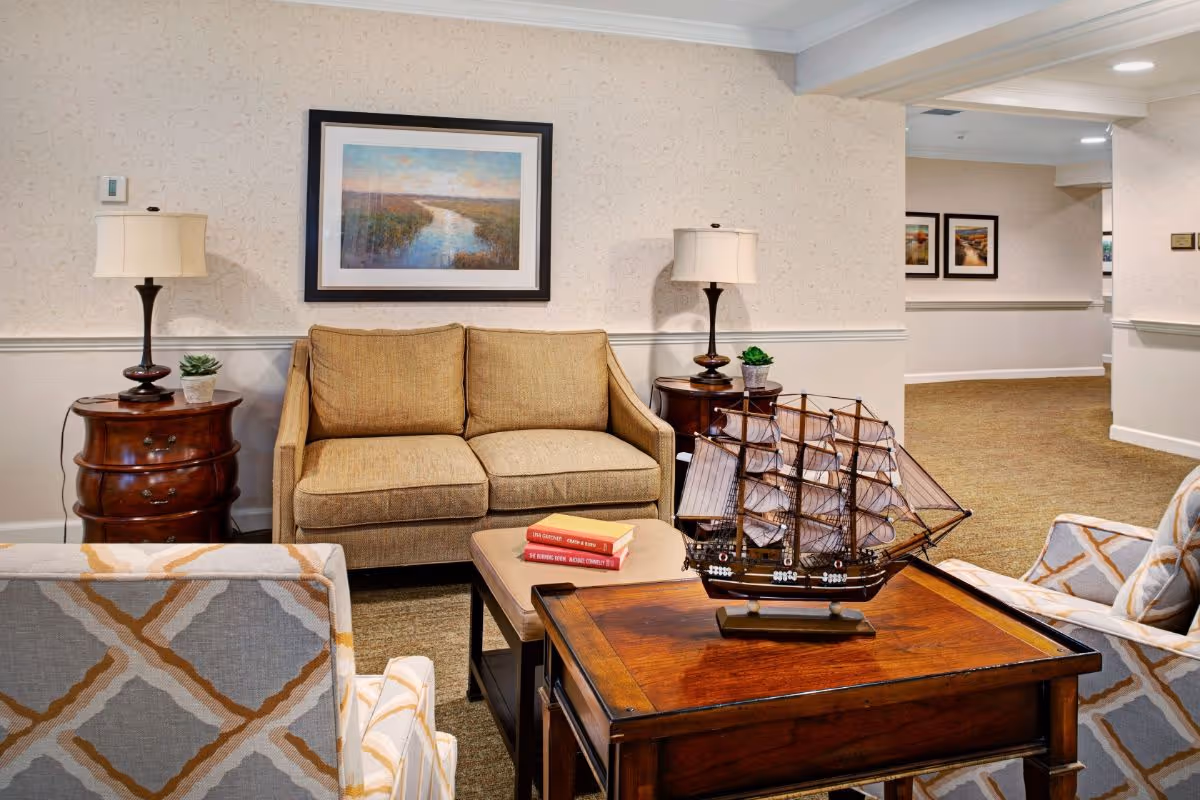 A cozy living room area with a beige loveseat, two patterned armchairs, and a wooden coffee table featuring a model ship. Two side tables with lamps and small potted plants flank the loveseat. A framed landscape painting hangs on the wall above the loveseat. The room has beige walls and carpet, with a hallway visible in the background.