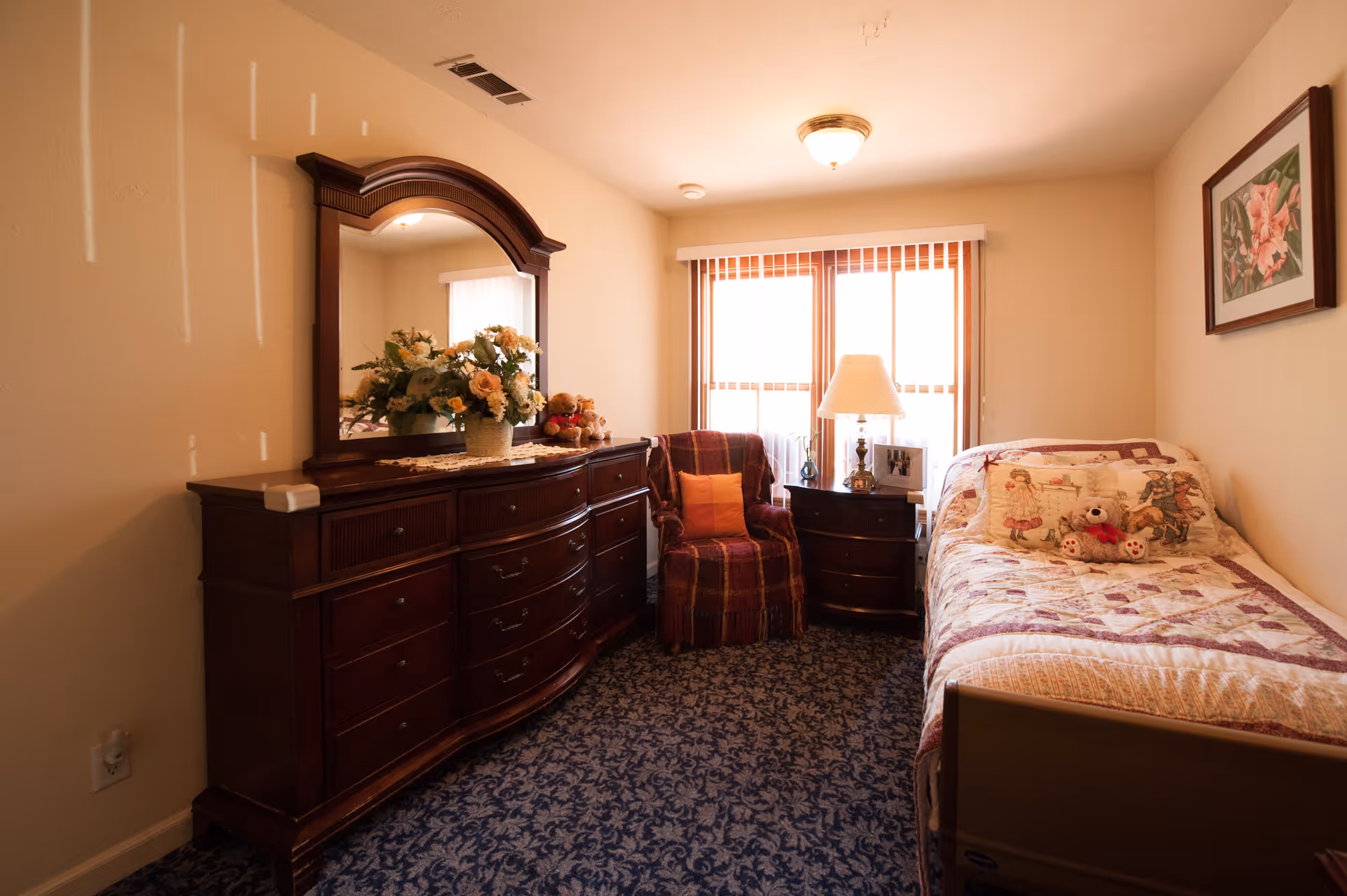 A cozy bedroom with a single bed covered in a patchwork quilt and decorative pillows, including a teddy bear. Next to the bed is a wooden nightstand with a lamp and framed photos. A comfortable armchair with a throw pillow is positioned near a window with vertical blinds. On the opposite wall, there is a large wooden dresser with an ornate mirror and a floral arrangement on top. The room has patterned carpet and a framed floral picture on the wall.