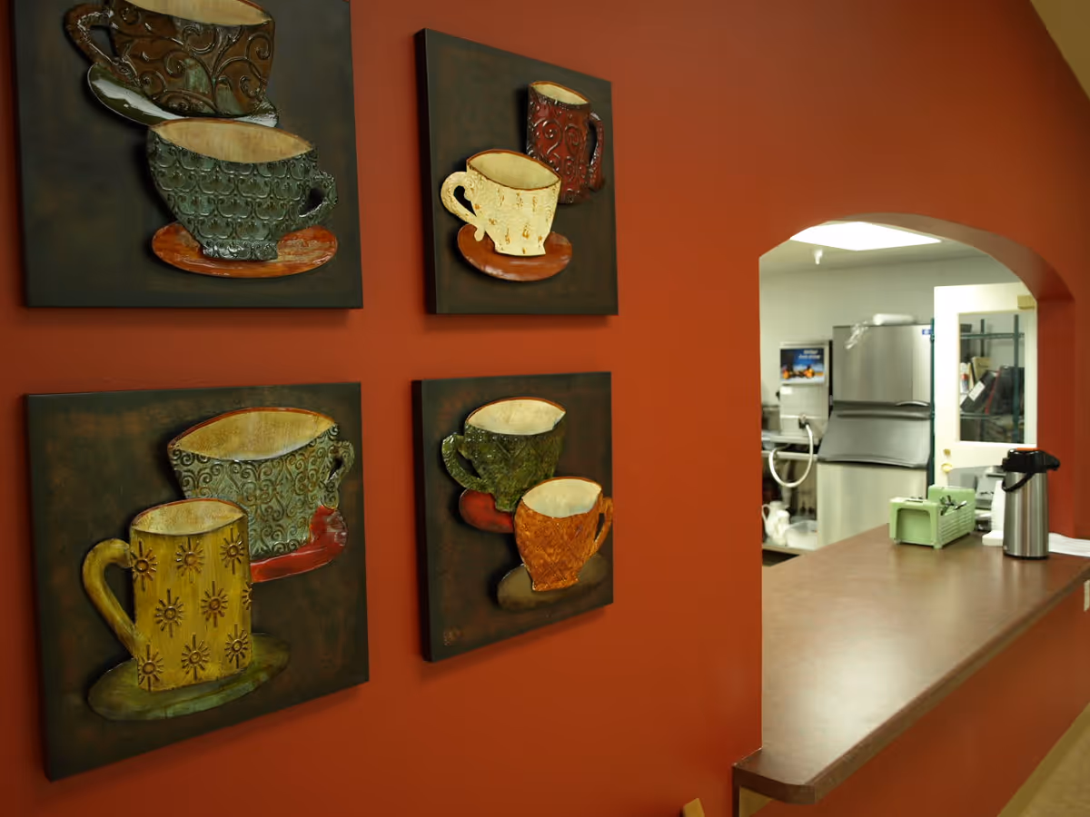 A red wall decorated with four square art pieces featuring colorful coffee cups and saucers. To the right, there is a counter with a coffee pot and a green napkin holder, and an open window looking into a kitchen area with stainless steel appliances.