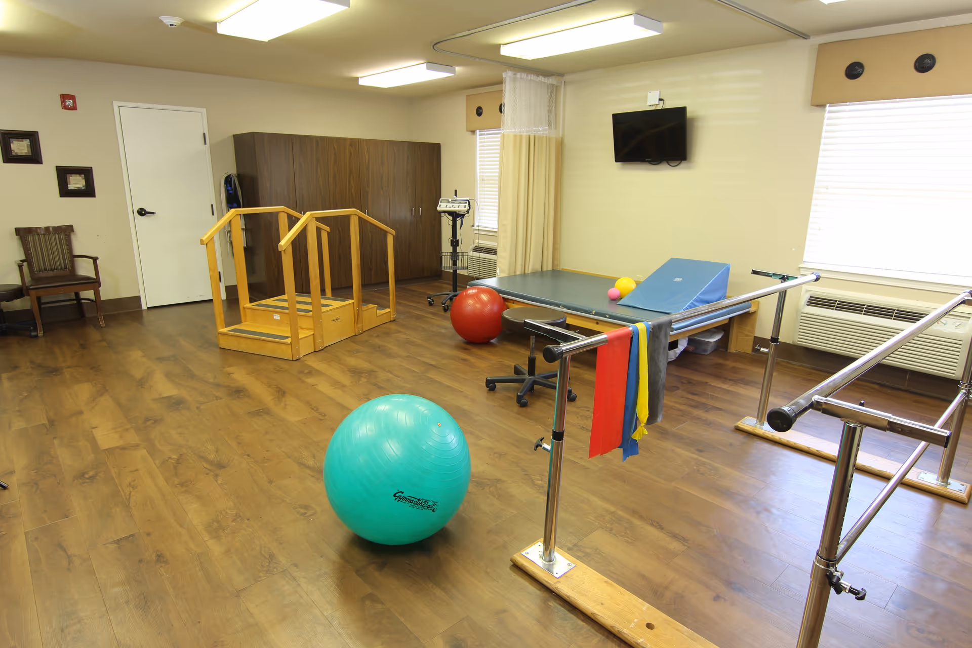 A physical therapy room with wooden flooring, featuring parallel bars for walking exercises, a large green exercise ball, a red exercise ball, a padded therapy table with a blue wedge cushion, resistance bands hanging on the parallel bars, a wooden step platform with handrails, a wall-mounted TV, and a wooden chair near a closed white door.