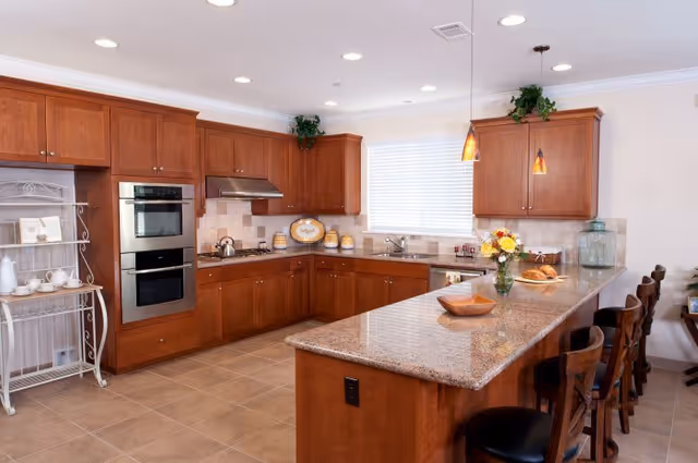 A spacious kitchen with wooden cabinets, granite countertops, and tiled flooring. The kitchen features a built-in double oven, a stovetop with a range hood, a sink under a window with blinds, and a large island with bar stools. There are decorative items such as a vase with flowers, bowls, and jars on the countertops. Pendant lights hang above the island.