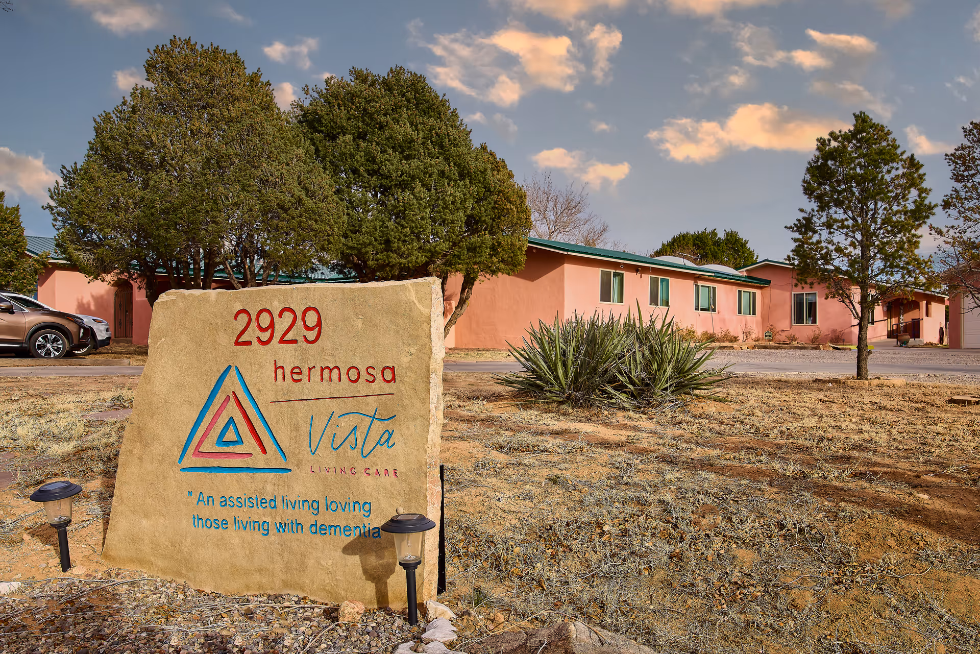 Stone entrance sign reading '2929 hermosa Vista' in front of a pink one-story senior living building with trees and dry landscaping.