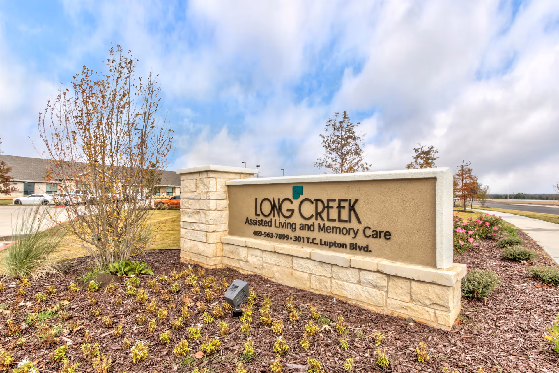Outdoor view of a stone and stucco sign for Long Creek Assisted Living and Memory Care, surrounded by landscaped plants and trees with a partly cloudy sky in the background.