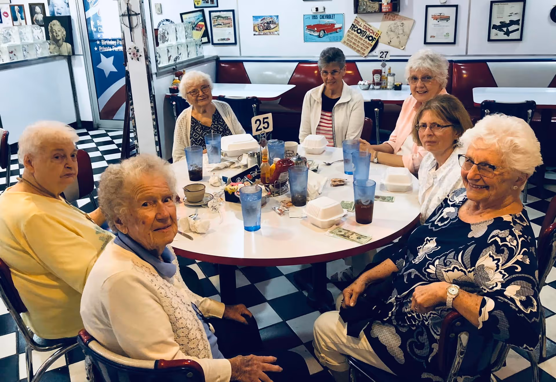 A group of seven elderly women sitting around a round table in a retro-style diner with checkered black and white floor tiles. The table has several blue and clear drinking glasses, takeout containers, condiments, and a table number 25. The walls are decorated with vintage posters and pictures.