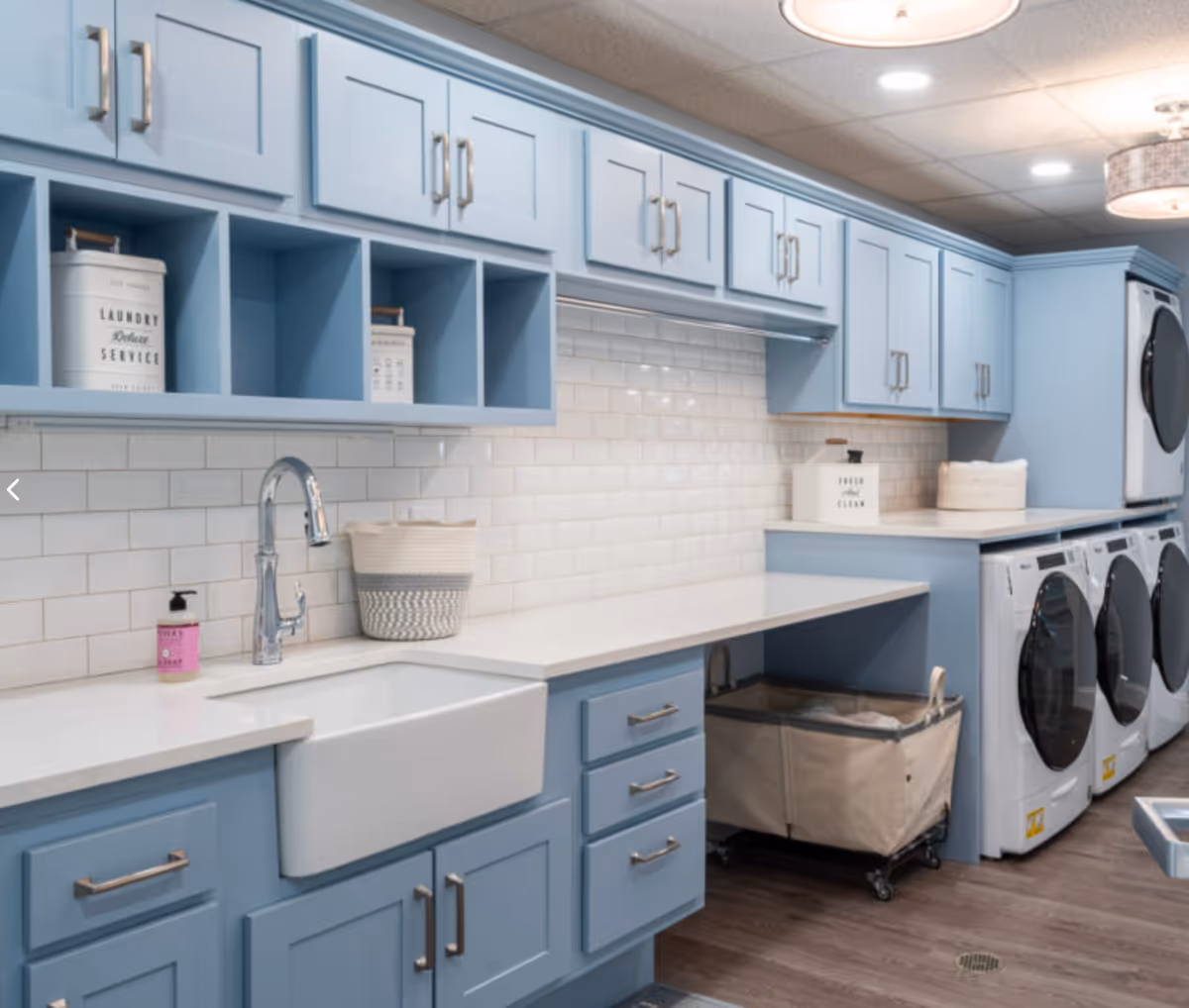 Laundry room with light blue cabinets, a white farmhouse sink, countertop workspace, and stacked front-loading washers and dryers.