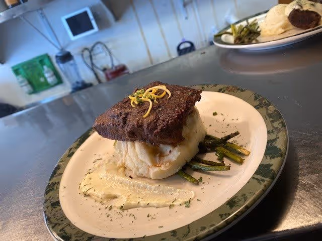 A plated meal featuring a piece of cooked steak topped with shredded cheese, served on a bed of mashed potatoes and accompanied by cooked asparagus, with a creamy sauce spread on the side of the plate. The plate is on a stainless steel countertop in a kitchen setting.