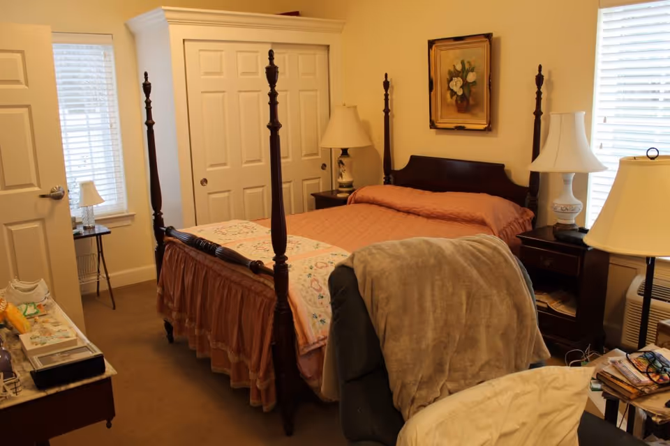 A cozy bedroom in an assisted living facility featuring a four-poster bed with a pink quilt and floral bedspread. The room has two windows with white blinds, two bedside tables each with a lamp, a recliner chair with a gray blanket draped over it, and a small table with books and glasses. There is a closet with white sliding doors and a framed floral painting on the wall.