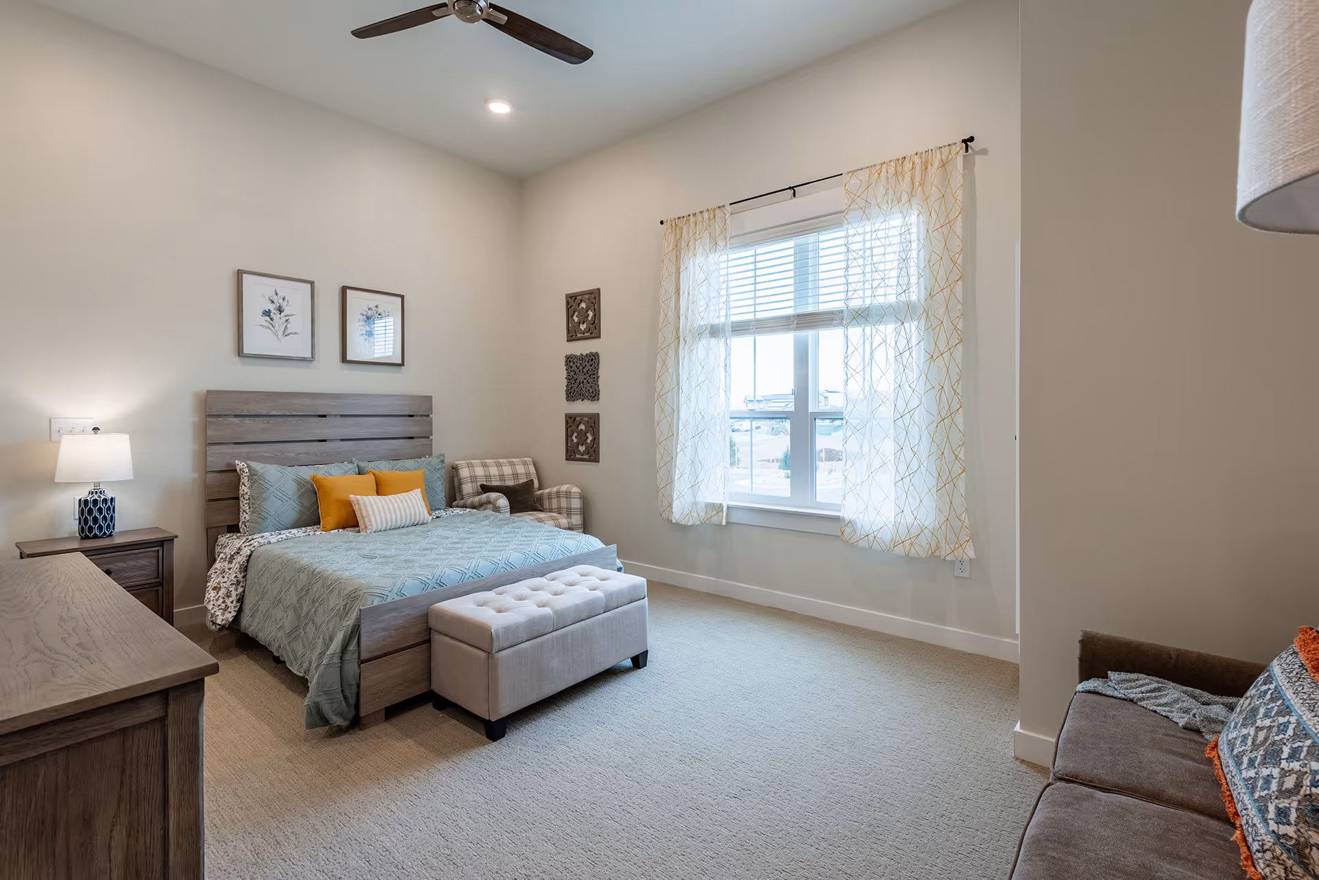 A bright bedroom with a wooden bed frame, light blue bedding, and several pillows in blue, yellow, and white. There is a small upholstered bench at the foot of the bed, a wooden nightstand with a lamp, and a wooden dresser. A window with sheer curtains lets in natural light. The walls are decorated with framed floral artwork and decorative wall hangings. A plaid armchair and part of a brown sofa with a patterned pillow are also visible.