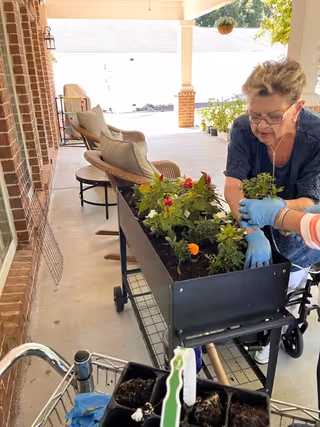 An elderly woman wearing blue gloves is gardening on a patio, planting flowers in a raised black planter. The patio has wicker chairs with cushions and a brick wall on one side. Another person, partially visible, is assisting her. There are potted plants and hanging planters in the background.