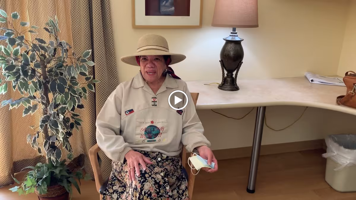 An elderly woman wearing a beige hat, a light-colored sweatshirt, and a floral skirt is sitting on a wooden chair in a room. She is holding a face mask in her right hand. Behind her is a large potted plant, a table with a lamp, some papers, and a brown bag. The room has beige walls and curtains.