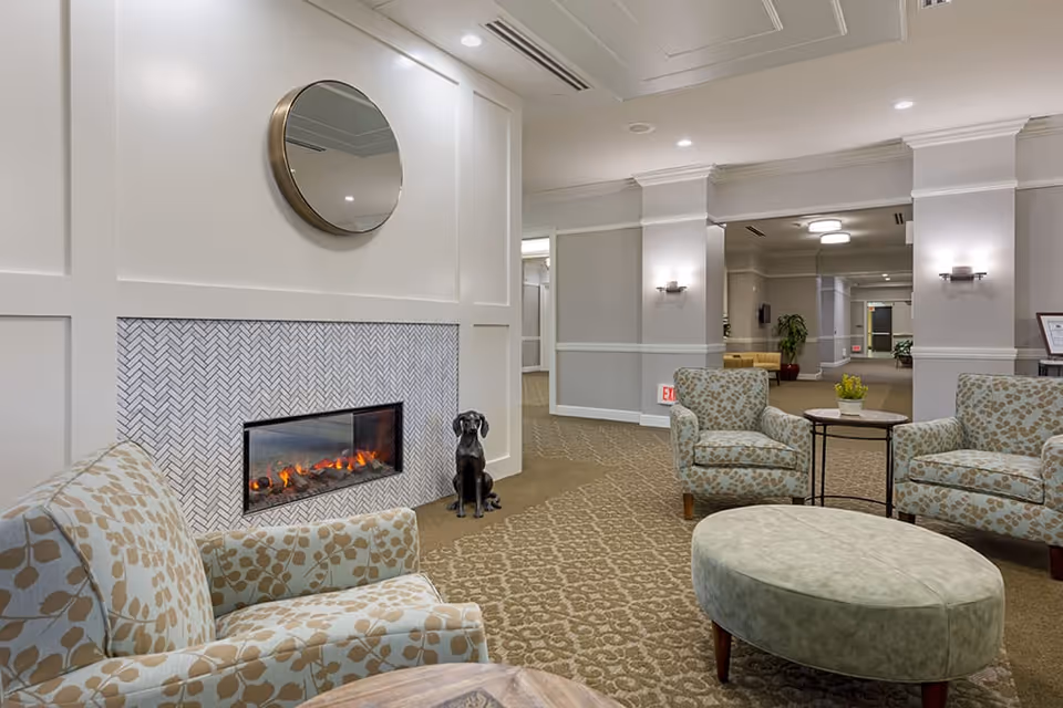 A cozy seating area in a senior living facility with patterned armchairs, a round ottoman, a small side table with a plant, a modern electric fireplace, and a round mirror above the fireplace. The space is well-lit with wall sconces and recessed ceiling lights, featuring neutral tones and carpeted flooring.