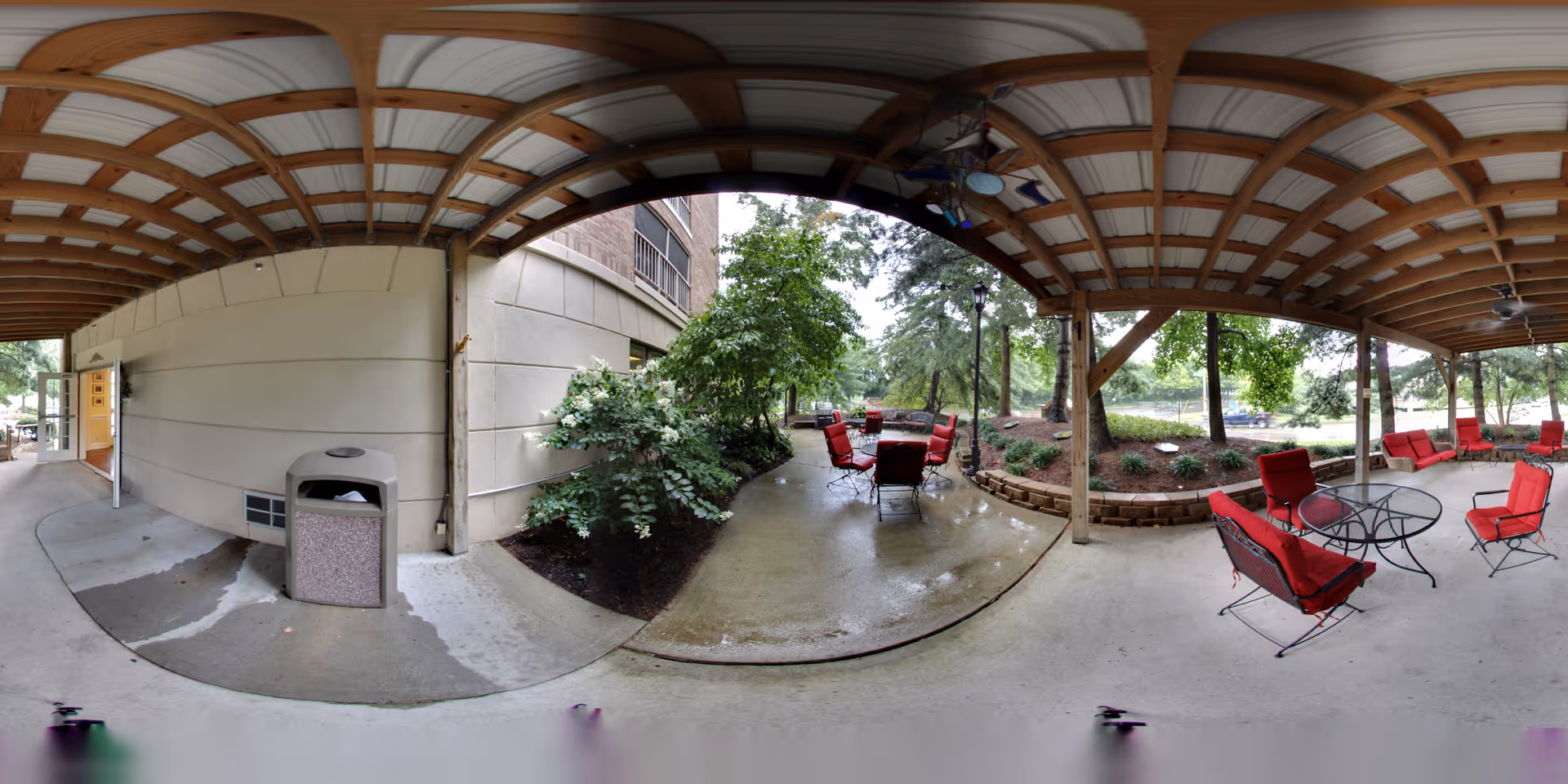 Covered outdoor patio area with a wooden beam ceiling and concrete floor. Several red cushioned chairs and black metal tables are arranged for seating. There are trees and landscaped garden beds surrounding the patio, with a building wall on one side and a trash can near the entrance.