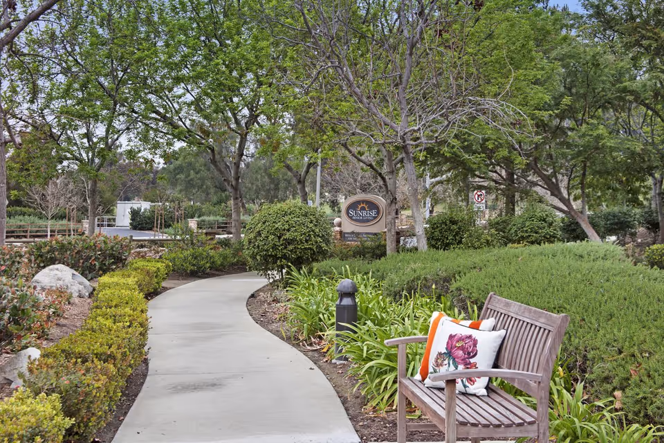 Curving paved walkway through landscaped grounds with a wooden bench and a 'Sunrise' community sign among trees.