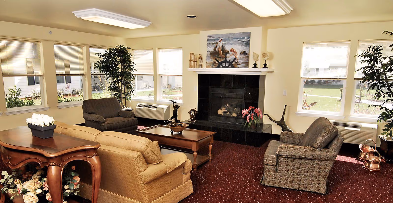 Bright communal living room with sofas and armchairs arranged around a coffee table and fireplace, with multiple windows letting in daylight.