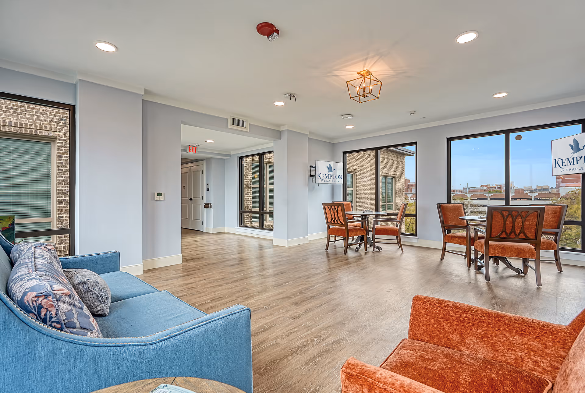 A bright and spacious common area in a senior living facility with large windows showing an outside view. The room features a blue sofa with patterned pillows, two orange upholstered chairs, and two small round tables with four chairs each. The walls are light-colored, and the floor is wood. There is a modern ceiling light fixture and a sign on the wall that reads 'Kempton of Charleston.'